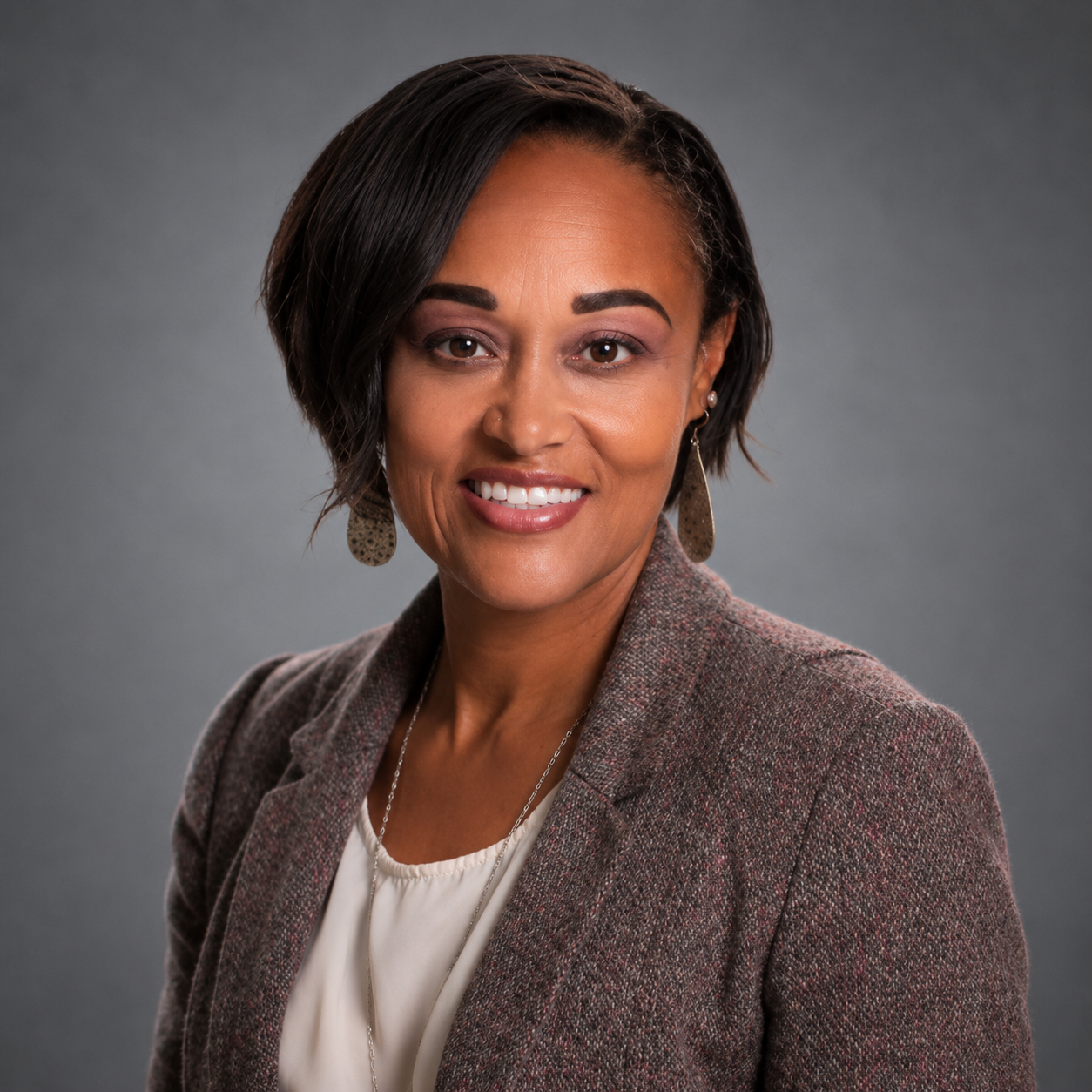 A woman with short dark hair is smiling and wearing a brown blazer and white shirt.